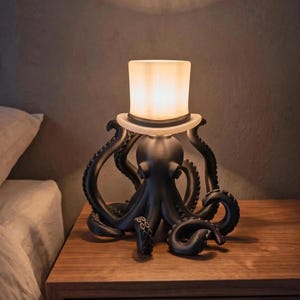 May include: A matte black octopus-shaped lamp base with multiple curved tentacles extending outward. The octopus wears a white, cylindrical top hat that functions as a glowing light diffuser, emitting a warm, soft light from within the structure.