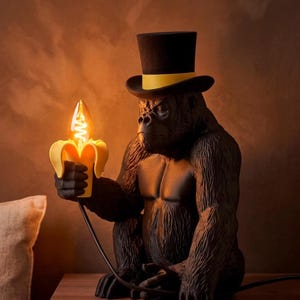 May include: A black gorilla figurine lamp wearing a black top hat with a yellow band. The gorilla holds a yellow banana-shaped light fixture with a glowing, flame-shaped light bulb. A black power cord extends from the banana light to the base.