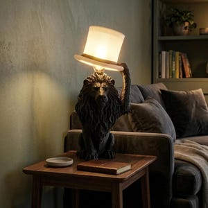 May include: A table lamp featuring a dark, textured lion sculpture in a seated position. The lion holds a white, cylindrical lampshade shaped like a top hat above its head. The lamp emits a warm, soft glow from beneath the hat-shaped shade.