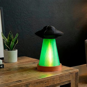 May include: A UFO-shaped lamp featuring a matte black top saucer section and a translucent green conical base that emits a bright light. The lamp rests on a circular terracotta-colored base, creating a distinct contrast between the dark top and light.