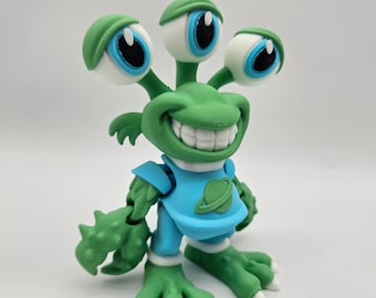Puede incluir: Una figura de alienígena verde de tres ojos con una amplia sonrisa de dientes. La criatura viste una camisa azul claro con un icono de planeta en relieve en el pecho. Tiene piel texturizada, manos en forma de garra y grandes ojos redondos.