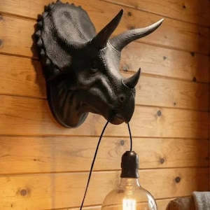 May include: A wall-mounted triceratops head sculpture in a matte black finish, featuring three horns and a textured frill. A black electrical cord hangs from the dinosaur's mouth, supporting a large, clear glass globe light bulb with a visible filament.
