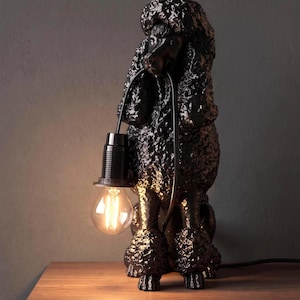 Könnte beinhalten: Eine schwarze Tischleuchte in Form eines Pudels mit strukturierter, gelockter Oberfläche. Die Hundefigur hält ein schwarzes Stromkabel im Maul, an dem eine einzelne, freiliegende Edison-Glühbirne mit schwarzer Fassung nach vorne hängt.