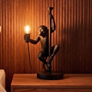 May include: A black table lamp featuring a sculpted monkey figure climbing a vertical pole. The monkey holds a light bulb socket in one hand, extending it outward. The lamp has a circular base and a black power cord connected to the socket.