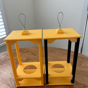 May include: Two yellow and black decorative stands with a square top and a circular opening in the center. Each stand has a wire loop handle attached to the top. The stands are made of plastic and sit on a wooden surface.