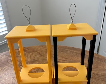 May include: Two yellow and black decorative stands with a square top and a circular opening in the center. Each stand has a wire loop handle attached to the top. The stands are made of plastic and sit on a wooden surface.