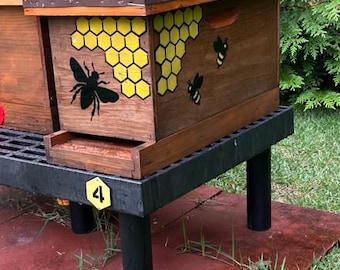 May include: A wooden beehive box featuring a painted yellow honeycomb pattern and black bee silhouettes on the side. The hive sits on a black plastic stand marked with a yellow hexagonal tag displaying the number 4, topped with a brown brick.