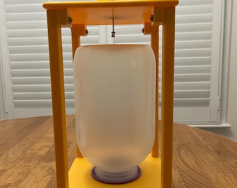 May include: A yellow lantern-style structure with a clear, cylindrical container inside. The lantern has a rectangular frame and a handle at the top. The container has a purple base and a black string attached to the top.