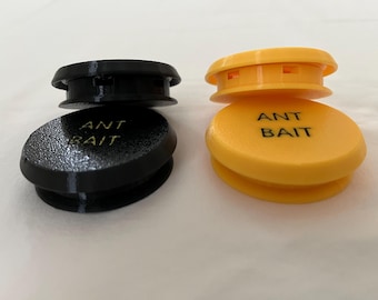 May include: Two ant bait stations, one black and one yellow. Each station has a circular base and a raised top. The text "ANT BAIT" is printed on the base of each station. The yellow station is stacked on top of the other.