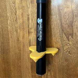 May include: A black GopherHawk device with a yellow base. The device has the words "Replaceable Straps" and "GopherHawk" printed on it. The device is designed to trap gophers.