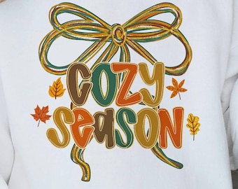 Puede incluir: Sudadera blanca con la frase "COZY SEASON" en colores otoñales. El diseño incluye un gran lazo y hojas. Una parte superior cómoda e informal para la temporada de otoño.