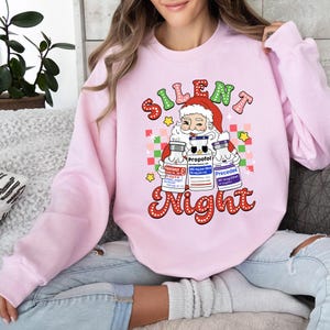 May include: A pale pink sweatshirt with a festive graphic. It features Santa Claus holding three medicine vials, with the words "Silent Night" in a decorative font. The design includes stars and a checkered pattern.