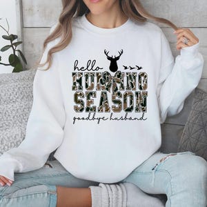 May include: White sweatshirt featuring the text "hello HUNTING SEASON goodbye husband". The words "HUNTING SEASON" are in a camouflage pattern. A black deer silhouette and three birds are above the text. The text is in black script and block letters.