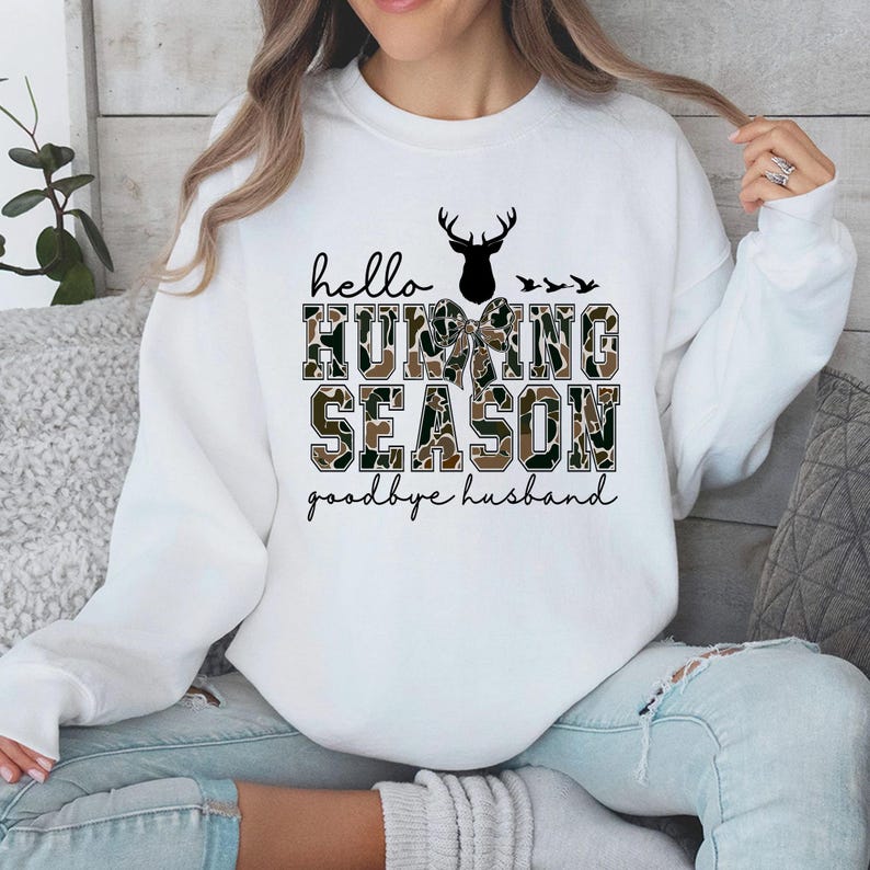 May include: White sweatshirt featuring the text "hello HUNTING SEASON goodbye husband". The words "HUNTING SEASON" are in a camouflage pattern. A black deer silhouette and three birds are above the text. The text is in black script and block letters.