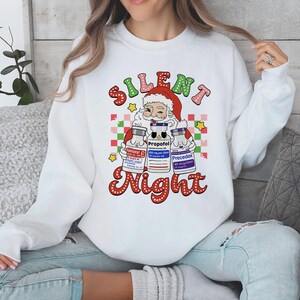 May include: White sweatshirt with a graphic of Santa Claus holding three medicine vials. The text reads "Silent Night" in a retro font. The vials are labelled "Fentanyl", "Propofol", and "Precedex".