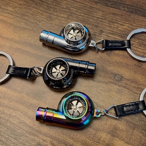 May include: Three turbocharger keyrings in silver, black, and iridescent finishes. Each keyring features a detailed turbine design and a black leather strap attached to a metal ring. The keyrings are displayed on a wooden surface.