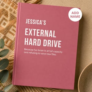 May include: A dusty rose hardcover book with white text that reads JESSICA'S EXTERNAL HARD DRIVE. Below this, smaller text reads: Because her brain is at full capacity and refusing to save new files. A white circular sticker says ADD NAME.