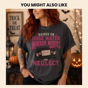 K&ouml;nnte beinhalten: Ein anthrazitfarbenes Kurzarm-T-Shirt mit einer Grafik einer schwarzen VHS-Kassette und tropfendem rosa Text, der lautet: RAISED ON HOSE WATER, HORROR MOVIES AND NEGLECT. Das Etikett in der Mitte der Kassette tr&auml;gt den Text SCARY MOVIE.