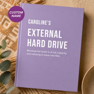 Könnte beinhalten: Ein lila Notizbuch mit dem weißen Text "CAROLINE'S EXTERNAL HARD DRIVE". Ein violetter Kreis oben links zeigt "CUSTOM NAME". Der untere Teil des Notizbuchs lautet "Because her brain is at full capacity and refusing to save new files."