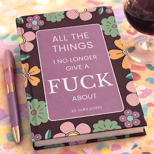 May include: A hardcover book with a floral design and the text "ALL THE THINGS I NO LONGER GIVE A FUCK ABOUT" by Sara Jones. A purple and gold pen rests beside the book. A glass of red wine is in the background.