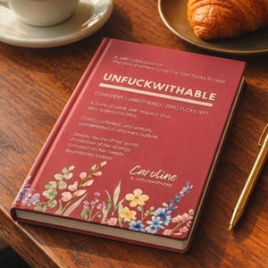 May include: A red self-care journal with the word "UNFUCKWITHABLE" in large white letters. The journal has floral designs and text that reads "Caroline is unfuckwithable". A gold pen, coffee cup, and croissant are in the background.