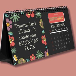 K&ouml;nnte beinhalten: Ein schwarzer Tischkalender mit Spiralbindung und Blumenmuster in Rot, Lila, Gr&uuml;n und Gelb. Der Text lautet: Trauma isn't all bad - it made you FUNNY AS FUCK. Ein rotes Feld enth&auml;lt den Text: Motto WHAT DOESN'T KILL YOU MAKES YOU HILARIOUS.