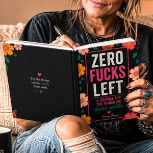 K&ouml;nnte beinhalten: Ein schwarzes Journal mit floralem Rand und dem Text Zero Fucks Left, A Journal for the Things I'm Done With von Sara Jones. Die linke Seite enth&auml;lt den Text For the things you're finally done with, akzentuiert durch kleine rosa Herzen.