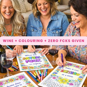 May include: Three coloring pages with floral borders and bold text. One page reads "What the ACTUAL FUCK on repeat." Another reads "Take it one SHITHOW at a time." The pages are surrounded by colorful markers and glasses of red wine on a wooden table.