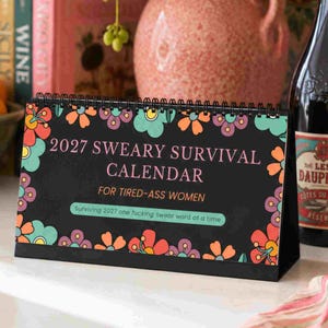 K&ouml;nnte beinhalten: Ein schwarzer Tischkalender mit Spiralbindung und buntem Blumenrand. Der Text lautet 2027 SWEARY SURVIVAL CALENDAR FOR TIRED-ASS WOMEN und Surviving 2027 one fucking swear word at a time in rosa, orangefarbener und hellgr&uuml;ner Schrift.