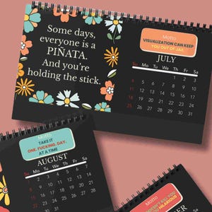 K&ouml;nnte beinhalten: Ein schwarzer Schreibtischkalender mit Spiralbindung und Blumenillustrationen in Orange, T&uuml;rkis und Wei&szlig;. Die Juli-Seite zeigt den Text 'Some days, everyone is a PI&Ntilde;ATA. And you're holding the stick.' sowie ein Motto-Feld mit 'VISUALIZATION CAN KEEP YOU OUT OF JAIL'.