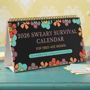 Può includere: Un calendario da scrivania con sfondo nero e bordo floreale. Il calendario recita "2026 SWEARY SURVIVAL CALENDAR FOR TIRED-ASS WOMEN" con il motto "Surviving 2026 one fucking swear word at a time."