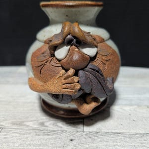 May include: A ceramic vase with a face design. The vase features a light beige body with brown and dark brown facial features, including eyes, a nose, and a mouth. The vase has a small opening at the top and a circular base.