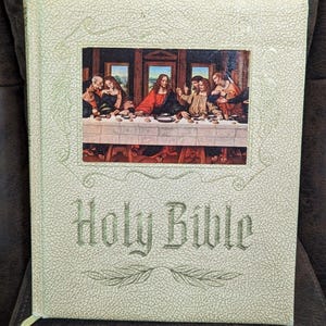 Könnte beinhalten: Eine gebundene Heilige Bibel mit einem strukturierten, cremefarbenen Einband. Die Vorderseite zeigt einen rechteckigen Einlegerdruck des Abendmahls mit Figuren an einem langen Tisch. Die Worte Holy Bible sind darunter in einer Schrift geprägt.