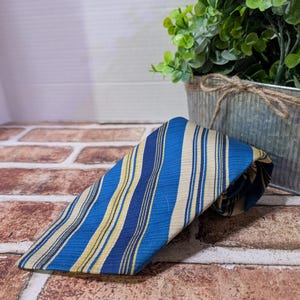 May include: A blue and yellow striped tie, angled on a brick surface. The tie features alternating vertical stripes of light blue, dark blue, and yellow. The tie is made of a textured fabric and is folded over itself.