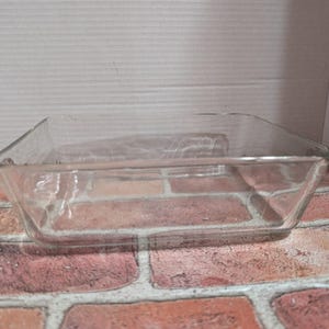 Puede incluir: Una fuente de horno cuadrada de vidrio transparente. La fuente tiene forma rectangular con esquinas ligeramente redondeadas y poca profundidad. Está sobre una superficie de ladrillo.