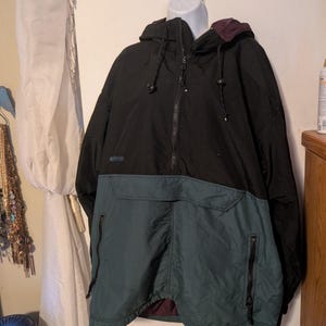 Könnte beinhalten: Schwarze und tealfarbene Anorakjacke mit Kapuze und Reißverschluss vorne. Die Jacke hat einen tealfarbenen unteren Teil mit zwei Reißverschlusstaschen und einen schwarzen oberen Teil. Die Jacke hängt an einem weißen Schaufensterkopf.