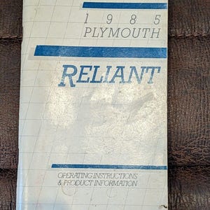 Puede incluir: Un folleto de instrucciones de funcionamiento e información de productos Plymouth Reliant de 1985. La portada es blanquecina con detalles azules y líneas de cuadrícula. El texto "RELIANT" destaca.