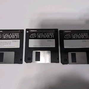 May include: Three black 3.5-inch floppy disks labeled "GALAXSEE SKYWATCH" and "DISK 1", "DISK 2", and "DISK 3". The disks have a silver sliding metal cover and white labels with black text. The brand "tasco" is printed at the top.