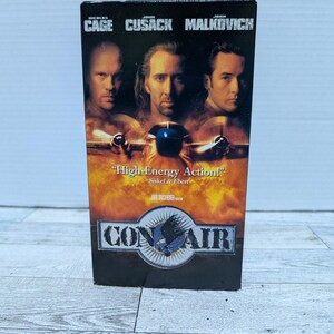 May include: A VHS tape for the movie "Con Air" featuring Nicolas Cage, John Cusack, and John Malkovich. The cover has a fiery background with the film's title and a quote from Siskel & Ebert.