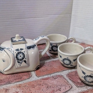 May include: A white ceramic teapot and three matching teacups. The teapot is shaped like an elephant with blue floral and geometric designs. The teacups also have blue floral patterns. The set is displayed on a brick surface.