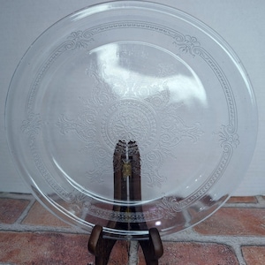 Peut inclure: Assiette en verre transparent avec un motif floral et volute orné. L'assiette est ronde et possède une bordure décorative. Le motif est gravé dans le verre, créant un aspect subtil et élégant. L'assiette est présentée sur un support en bois.