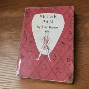 May include: A hardcover book titled Peter Pan by J. M. Barrie with a red cover featuring a white shield-shaped center panel. The cover displays a central illustration of Peter Pan surrounded by a diamond pattern containing small black line drawings.