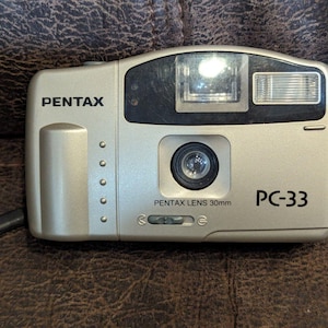 May include: A silver Pentax PC-33 camera with a 30mm lens. The camera has a built-in flash and a black wrist strap. The brand name "PENTAX" is printed on the top left, and "PC-33" is printed on the bottom right.