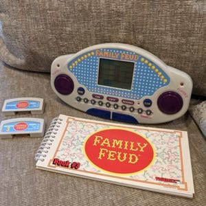 May include: A handheld Family Feud electronic game console with a blue and white polka dot face, two purple buttons, a digital screen, and number keys. Includes two grey game cartridges and a spiral-bound instruction booklet labeled Book #3.