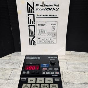 Puede incluir: Una caja de ritmos negra Zoom MRT-3B Micro RhythmTrak con una pantalla digital que muestra "180.1". El manual de instrucciones está detrás de la máquina, sujeto por un clip negro. El manual dice "Micro RhythmTrak ZOOM MRT-3 Operation Manual".