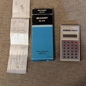 Puede incluir: Una calculadora Sharp EL-211 vintage con su caja original y manual de instrucciones. La calculadora es de color gris claro con botones rojos. La caja es negra y azul con el logotipo de Sharp y el número de modelo.
