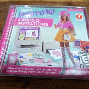 May include: A Barbie Print 'n Play CD-ROM case featuring a Barbie doll illustration. The case is pink and clear, with the words "CARDS & INVITATIONS" and "Barbie Print 'n Play" visible. The case also includes the ESRB rating.
