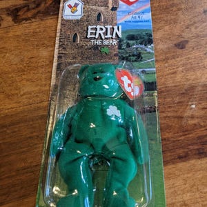 Puede incluir: Un oso de peluche verde llamado "Erin" con un trébol en el pecho, empaquetado en una funda de plástico transparente. El embalaje presenta el texto "Erin The Bear" y una ilustración de castillo. Una etiqueta roja en forma de corazón está adjunta.