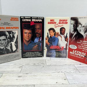 May include: Four VHS tapes of the "Lethal Weapon" movie series. The tapes feature Mel Gibson and Danny Glover. The first tape is gray, the second and third are black, and the fourth is red.