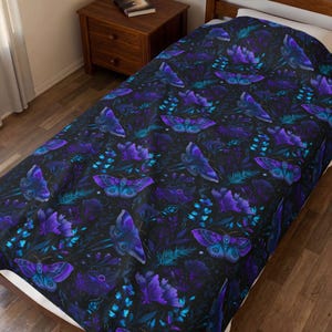 Op de afbeelding: Een donkere deken met een herhalend patroon van paarse en blauwe motten en bloemelementen. Het ontwerp bevat verschillende tinten paars, blauwgroen en blauw op een zwarte achtergrond. De deken ligt op een bed.